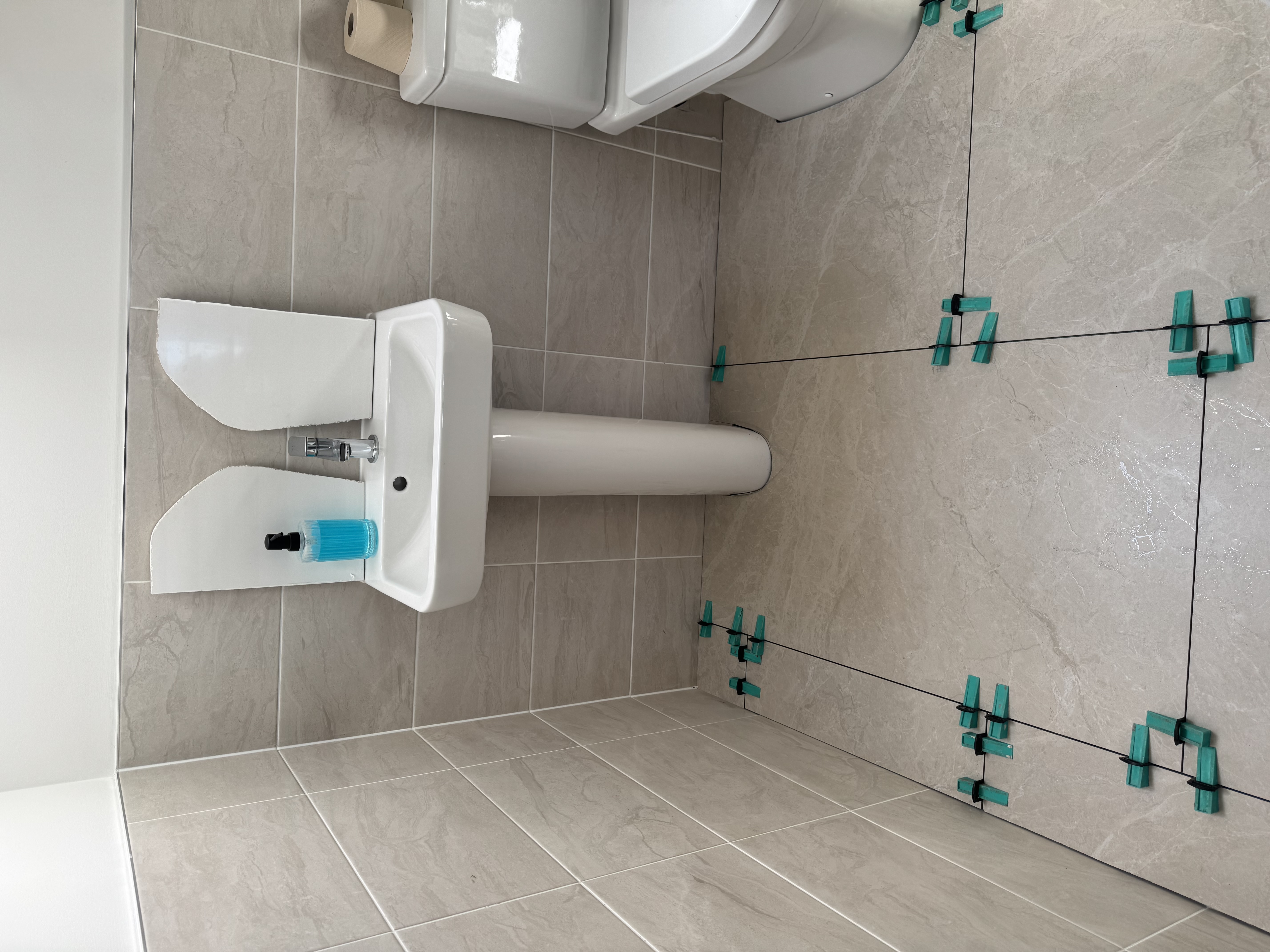 Newly tiled bathroom floor with levelling clips visible, light grey wall and floor tiles, a white pedestal sink, and a toilet.