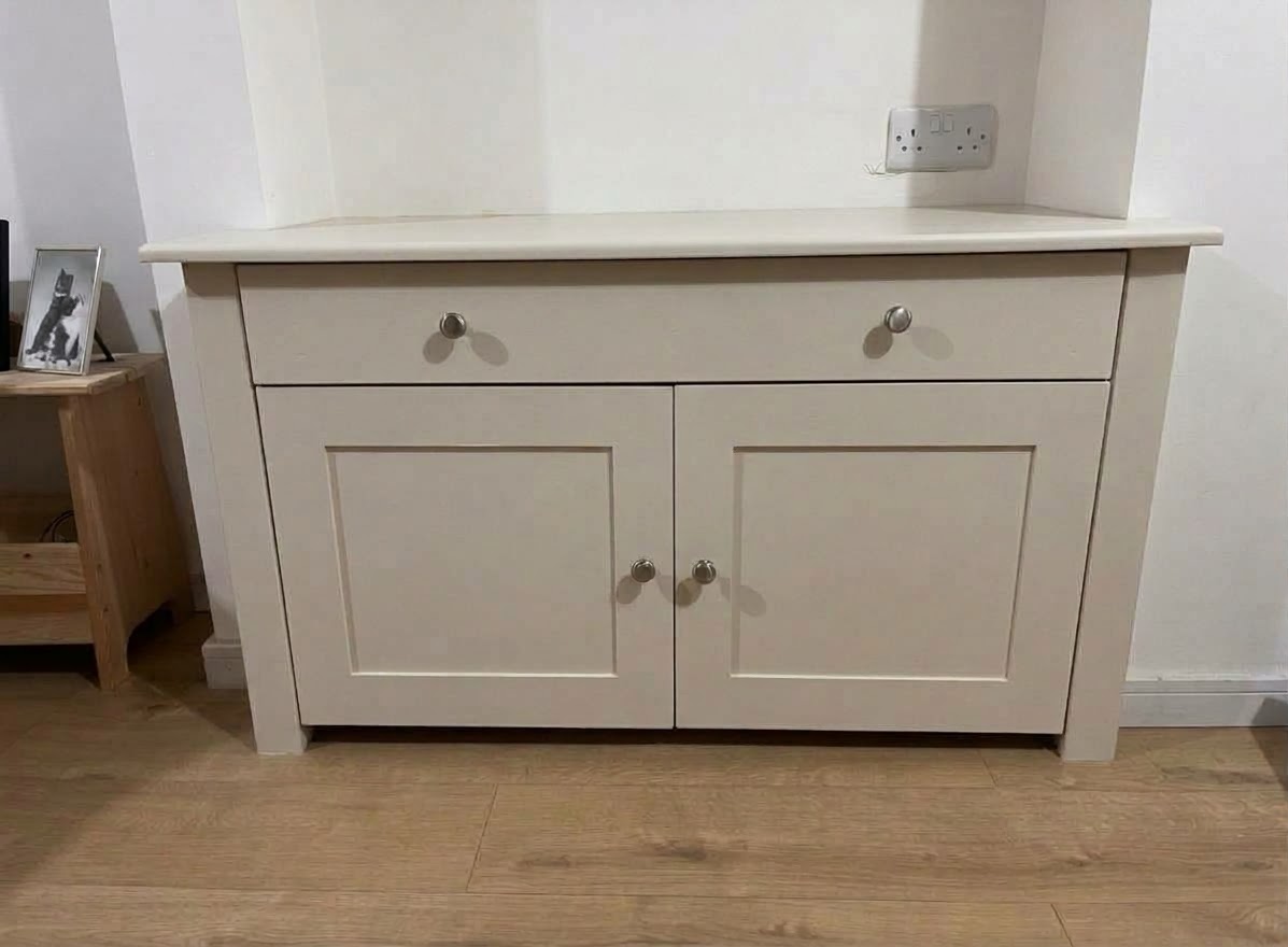 A fitted light grey wooden cabinet with two drawers and two doors, integrated into a wall recess with electrical sockets above.