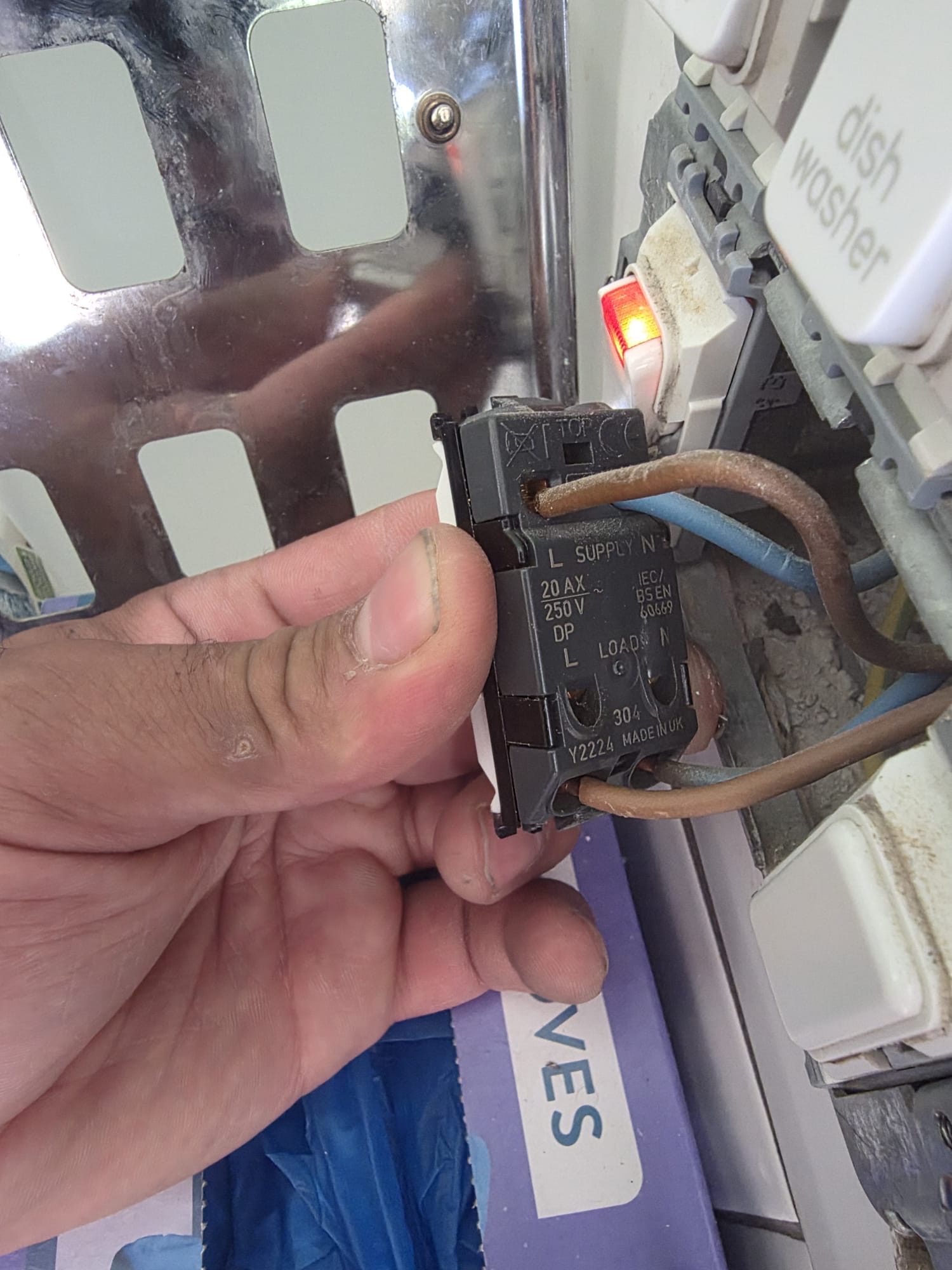 Close-up of a hand holding an electrical switch module with brown and blue wires, next to a white dishwasher switch.