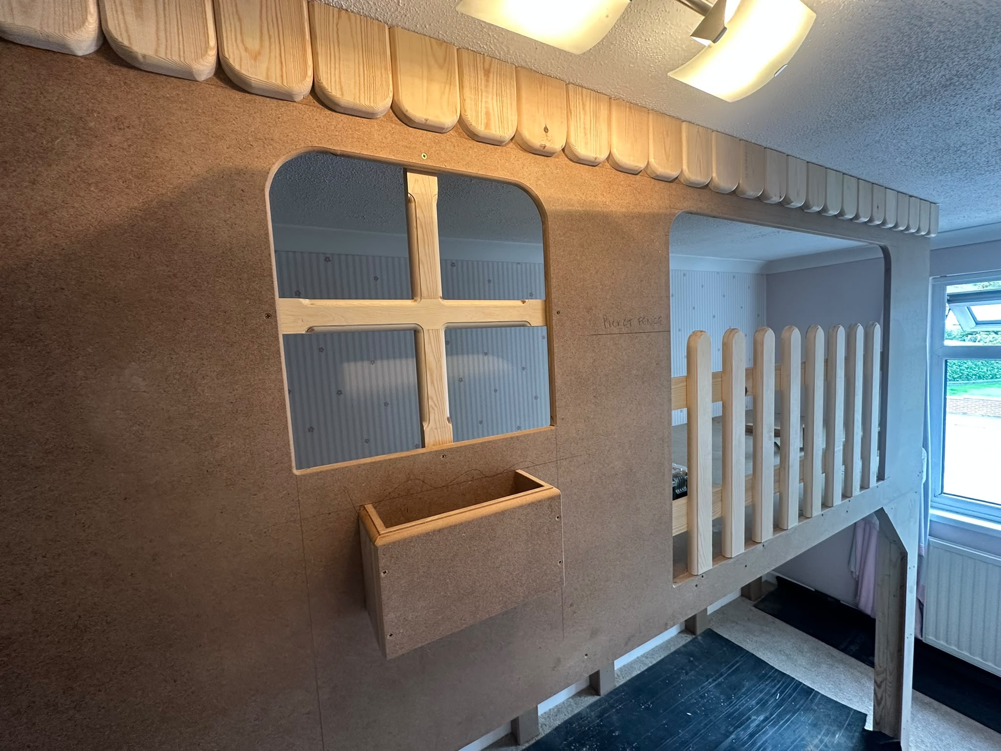 Unfinished custom-built MDF raised bed frame for a child's room, featuring a window, flower box, and picket fence railing.