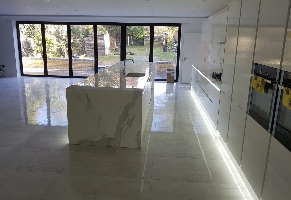 Sleek modern white kitchen with a large marble-effect island, integrated ovens, illuminated cabinets, and bifold doors to a garden.