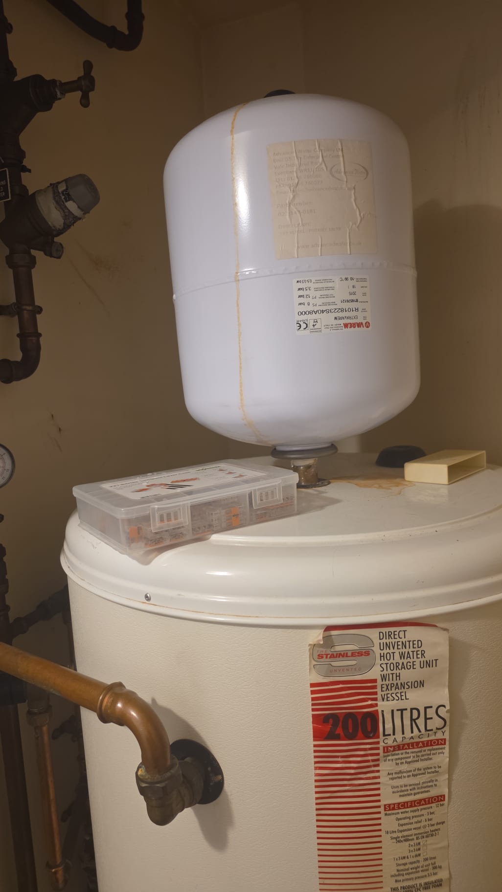 Unvented hot water storage cylinder with a white expansion vessel, copper pipes, and control valves in a utility space.