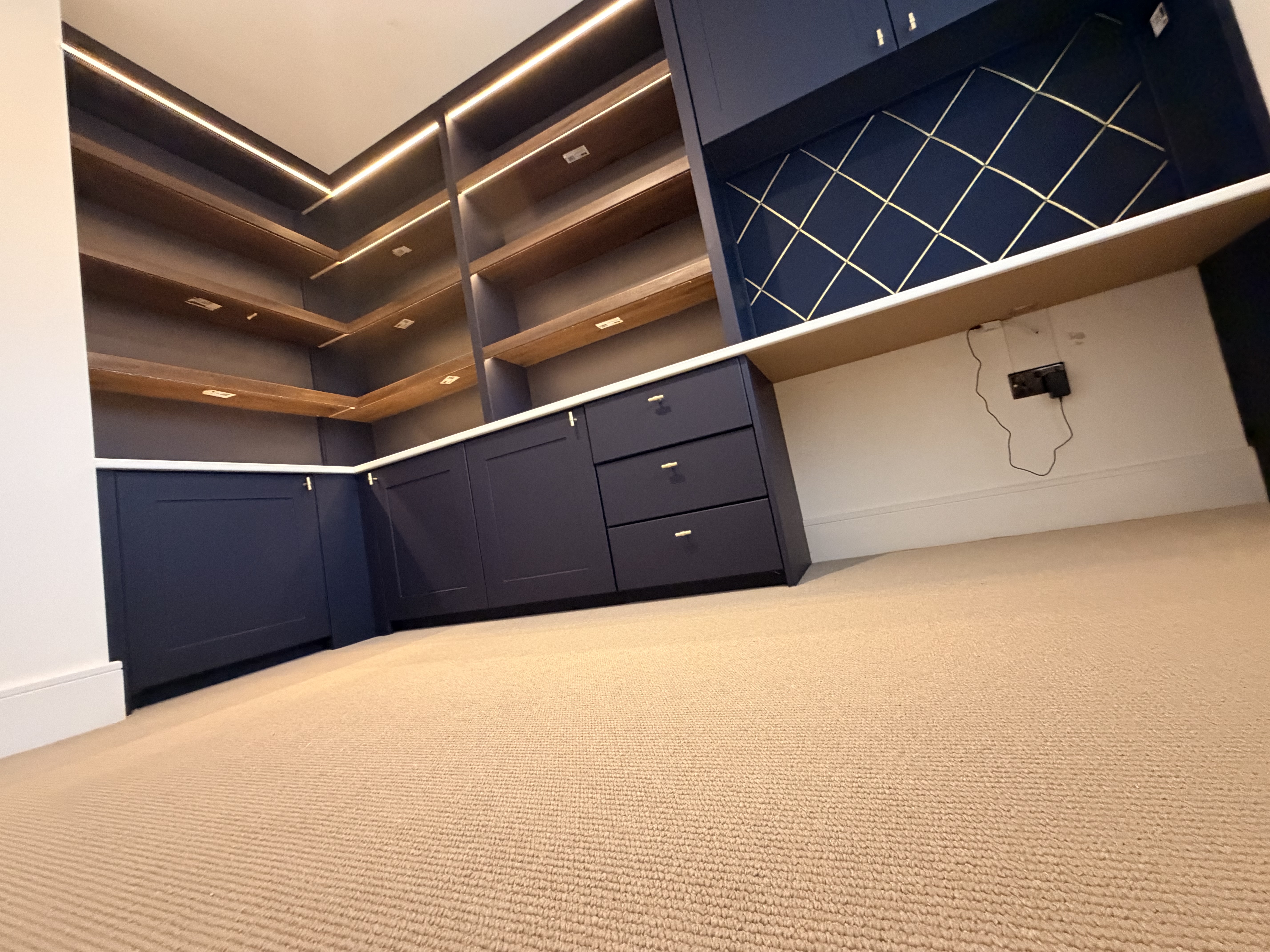 Dark blue bespoke fitted cabinetry with wooden shelves and integrated lighting in a home office, featuring a carpeted floor.