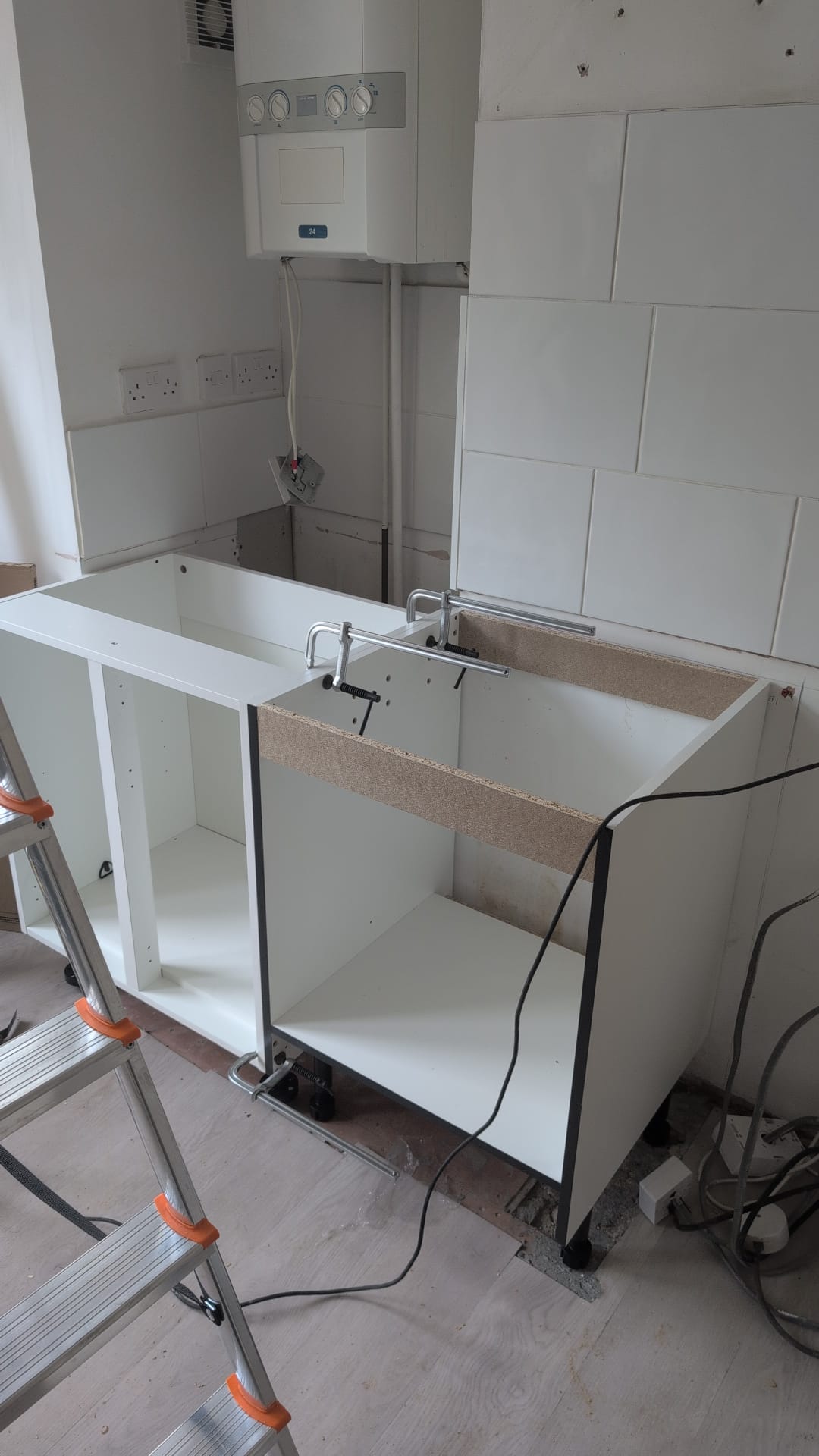 Installation of white kitchen base cabinets in a home with a boiler, wall tiles, and electrical sockets. Work in progress.