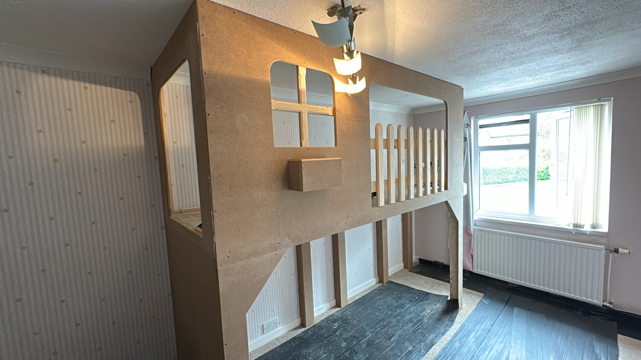 Unfinished custom-built MDF playhouse or raised bed structure in a room, showing raw wood panels and a small picket fence.