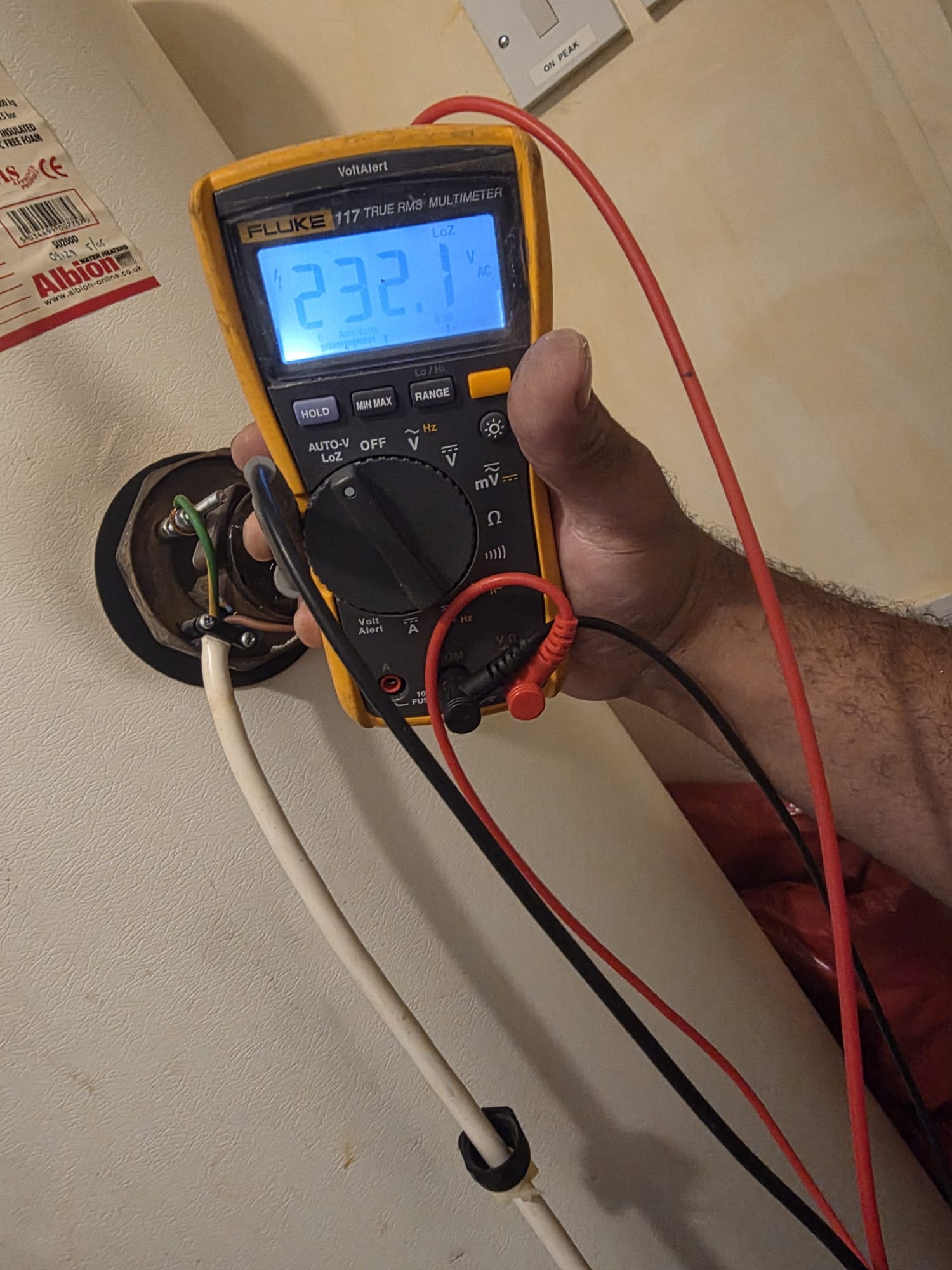 Electrician's hand holding a Fluke multimeter, displaying 232.1 V AC during electrical testing of wiring for a hot water cylinder.