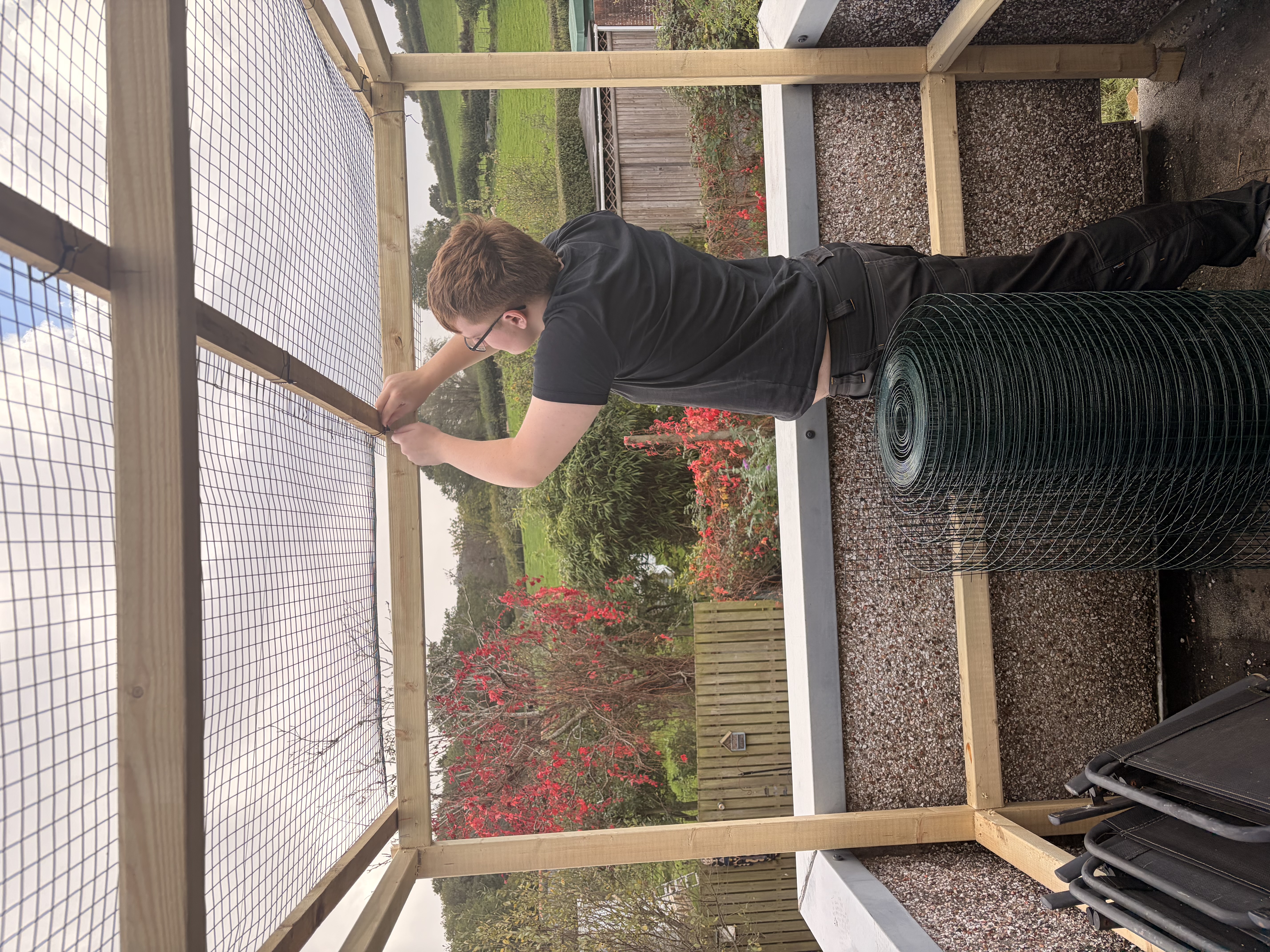 Tradesperson installing wire mesh on a new wooden frame structure on an outdoor balcony or patio with rural background.