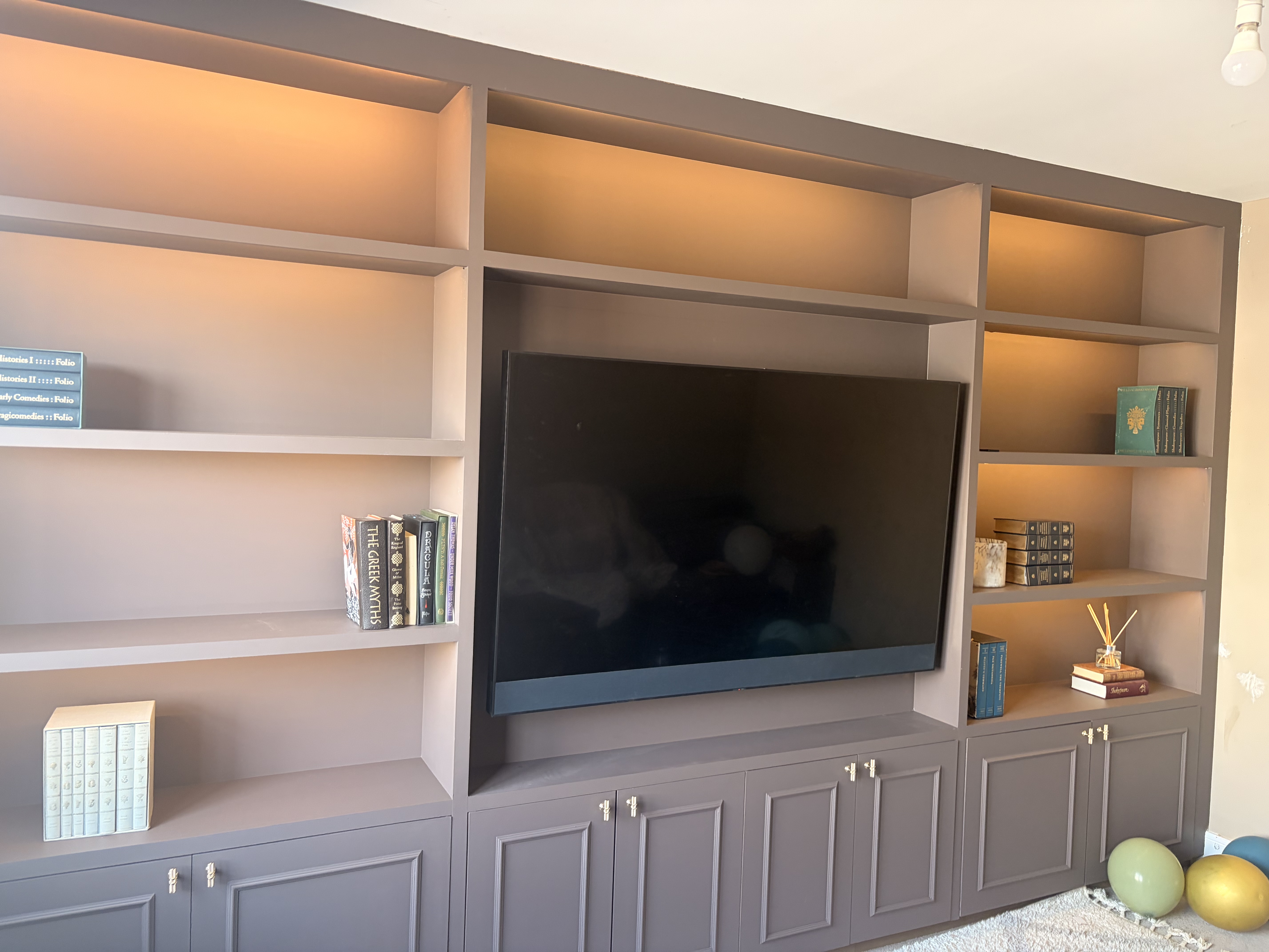 Custom-built grey-painted wall unit with open bookshelves, integrated lighting, a central TV recess, and lower storage cabinets.