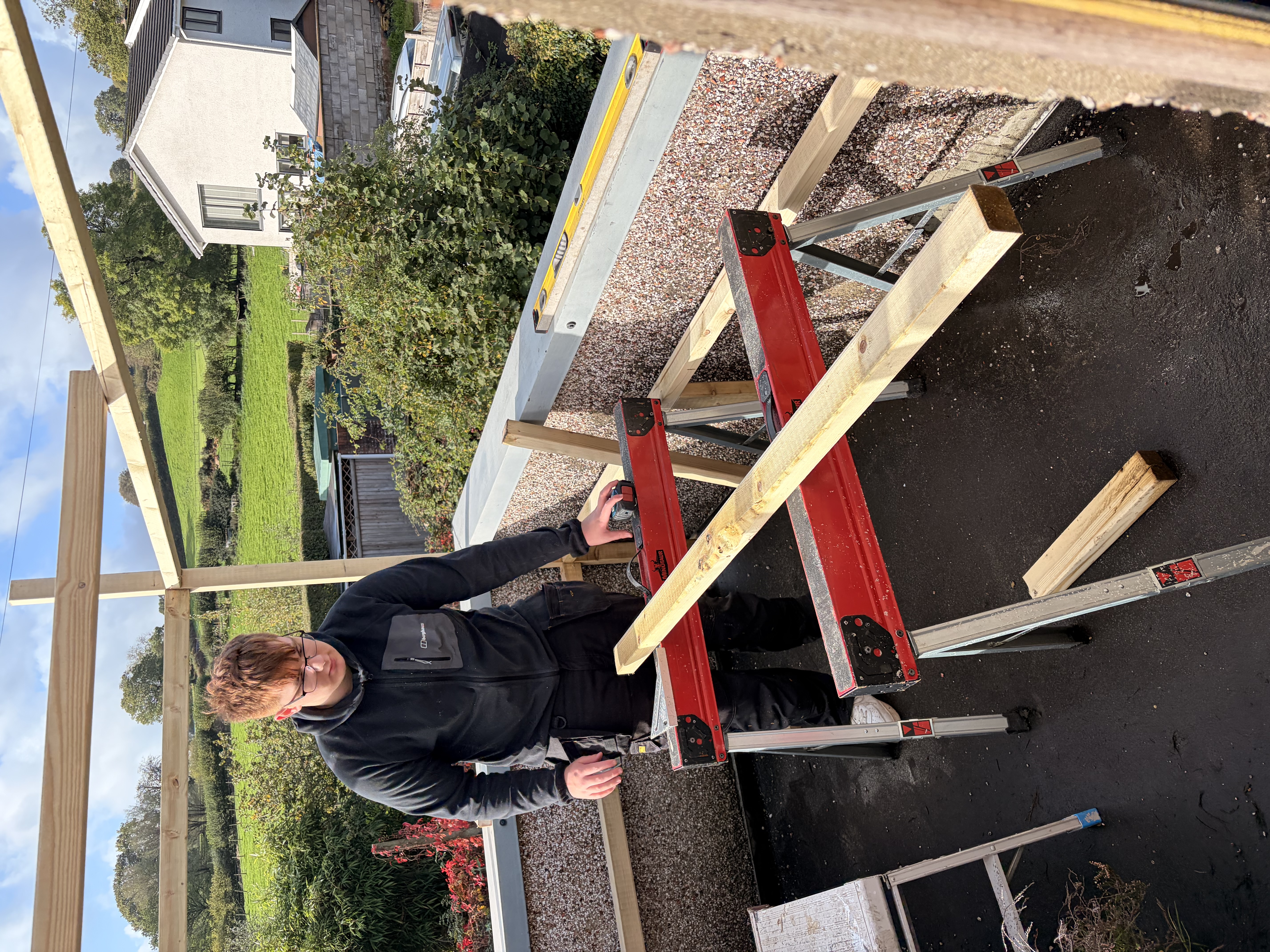 A tradesperson in black workwear cuts timber on red saw horses, constructing an outdoor timber frame structure on a balcony.