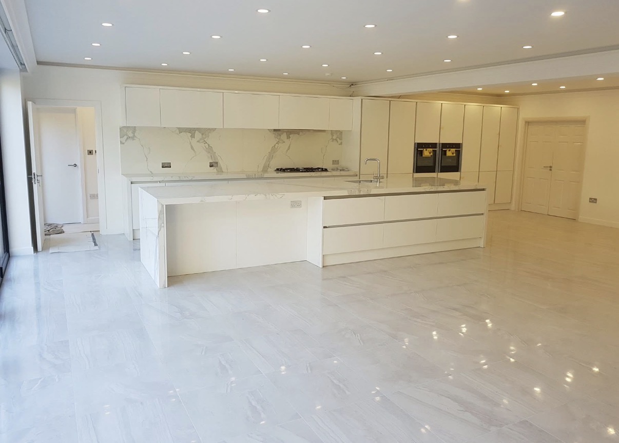 A spacious modern white kitchen featuring a large island, marble-effect countertops and splashback, integrated appliances, and light grey tiled flooring.