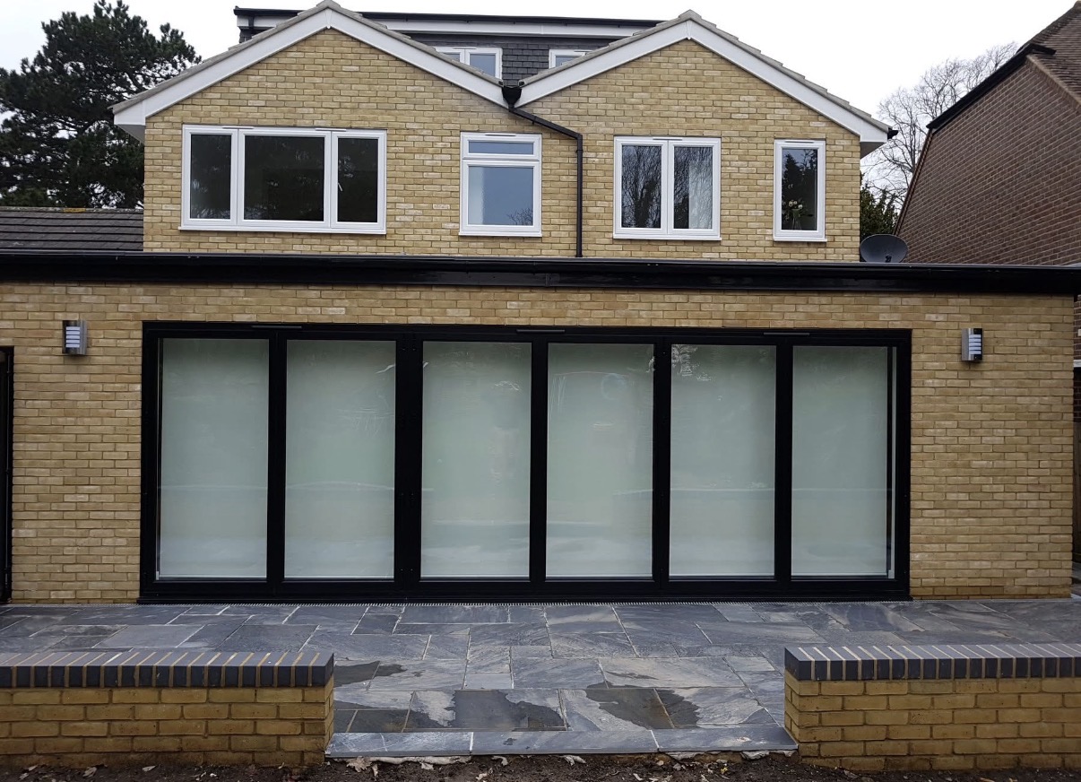 Modern house extension featuring large black bi-fold doors, new light yellow brickwork, white windows upstairs, and a dark paved patio.