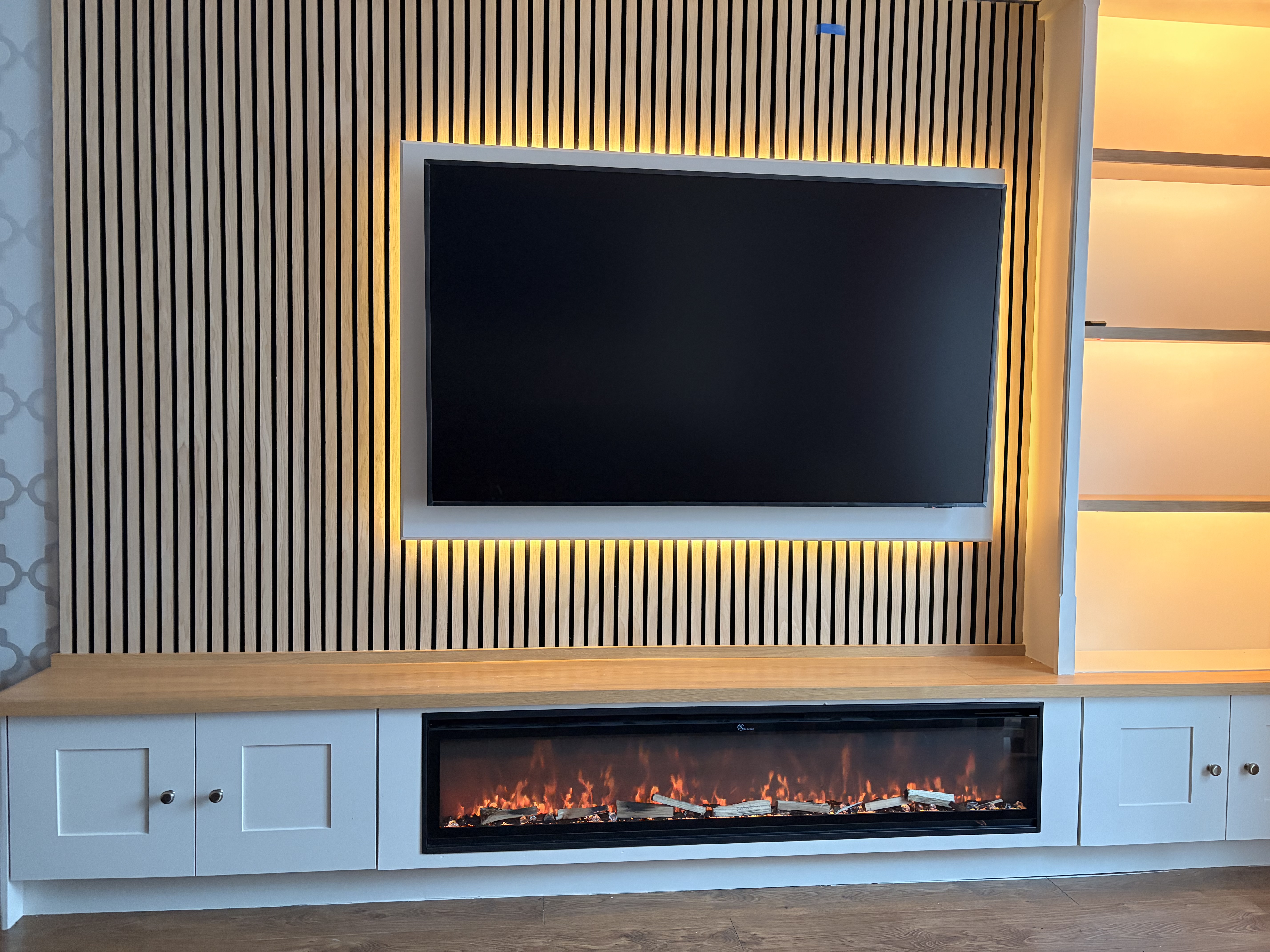 Bespoke carpentry media unit with wooden slat wall, integrated TV, electric fireplace, white cabinets, and illuminated shelving.