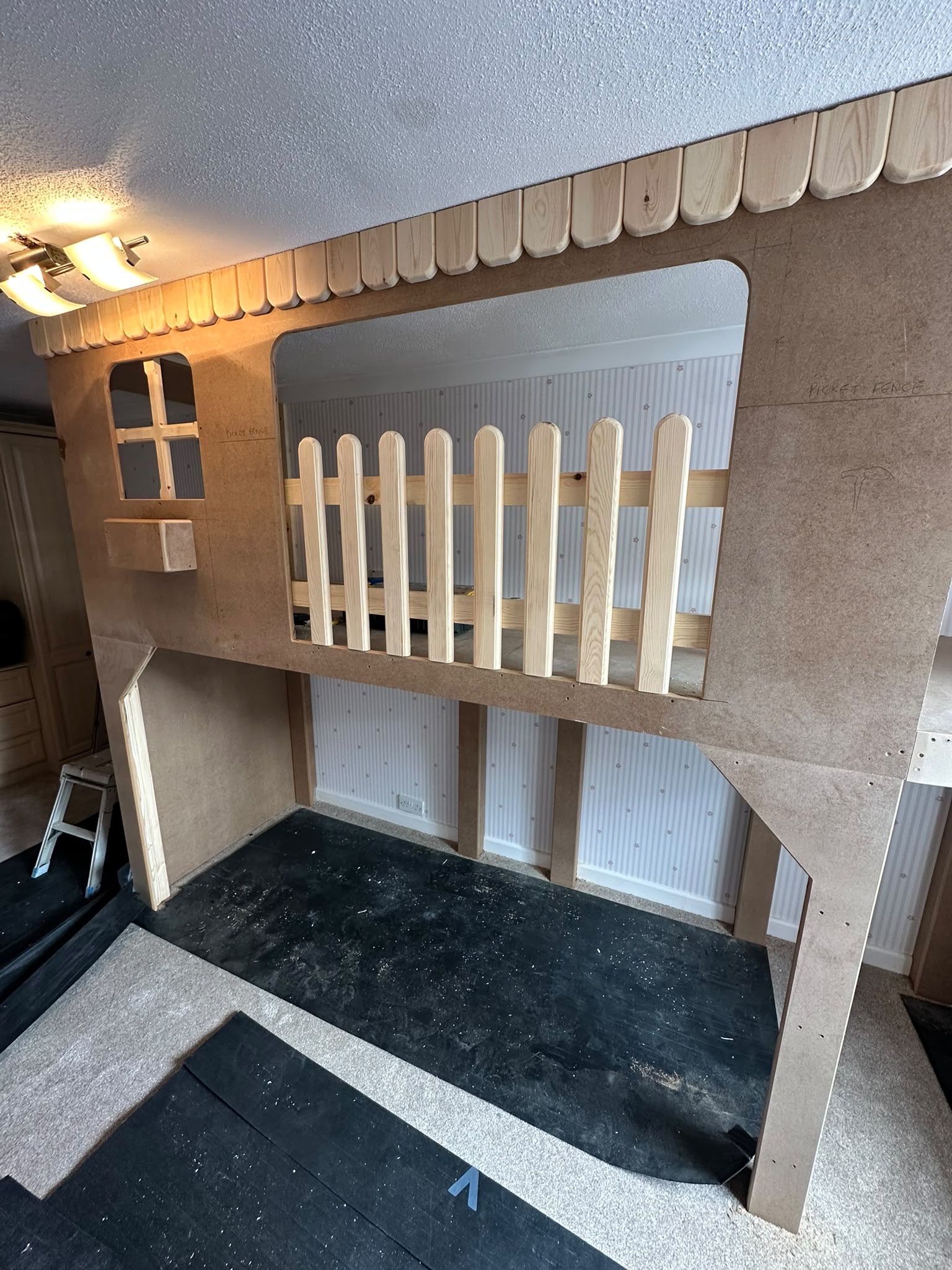 Partially built house-shaped loft bed frame with a picket fence design, constructed from MDF and wood in a child's bedroom.