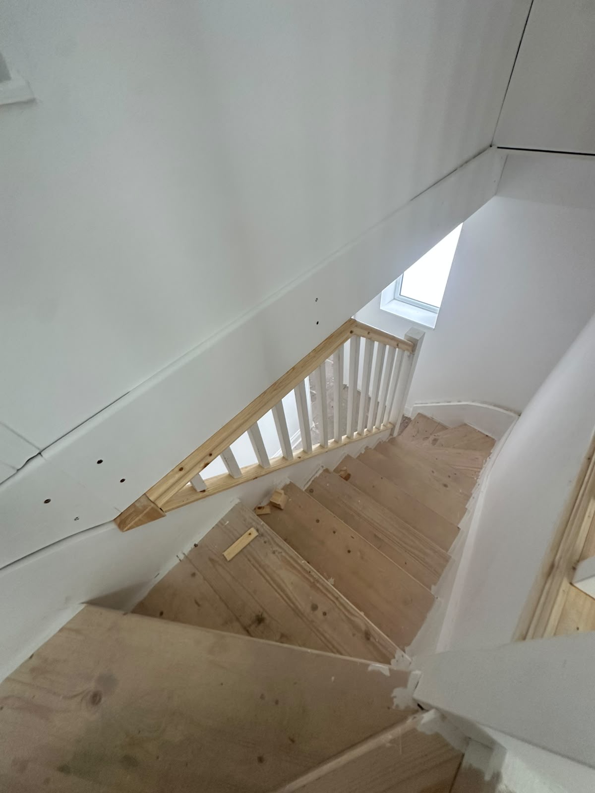 Unfinished wooden staircase with new wooden banister and white spindles, leading up to a skylight in a newly renovated space.