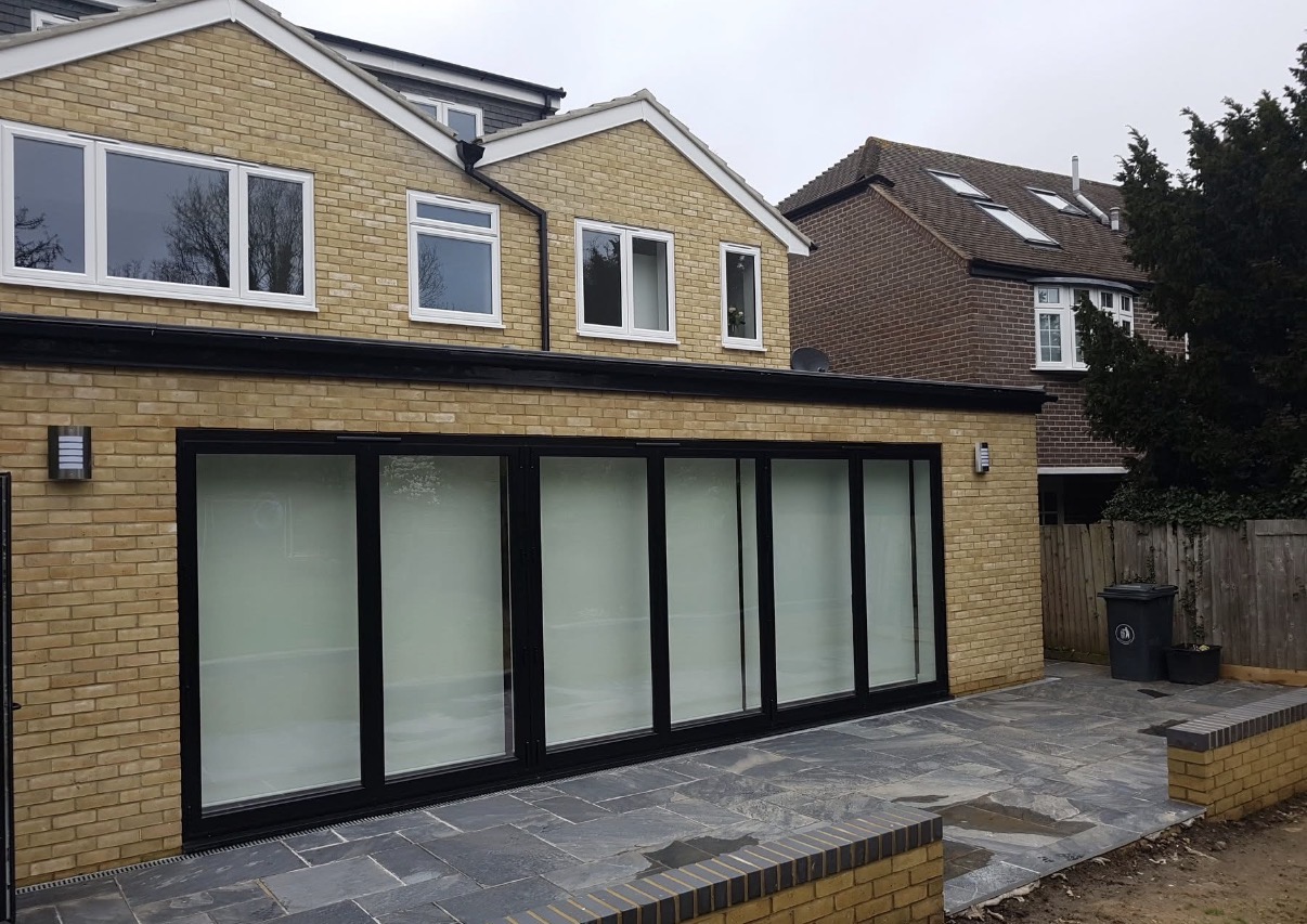 Exterior view of a house extension featuring yellow brickwork, black bi-fold doors, and a grey flagstone patio area.