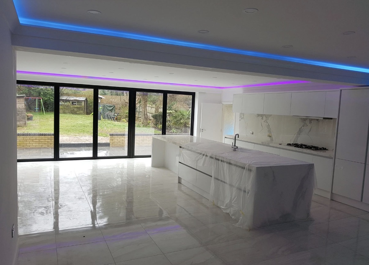 Modern white kitchen with a large island, tiled floor, recessed and strip lighting, and bi-fold doors opening to a garden.