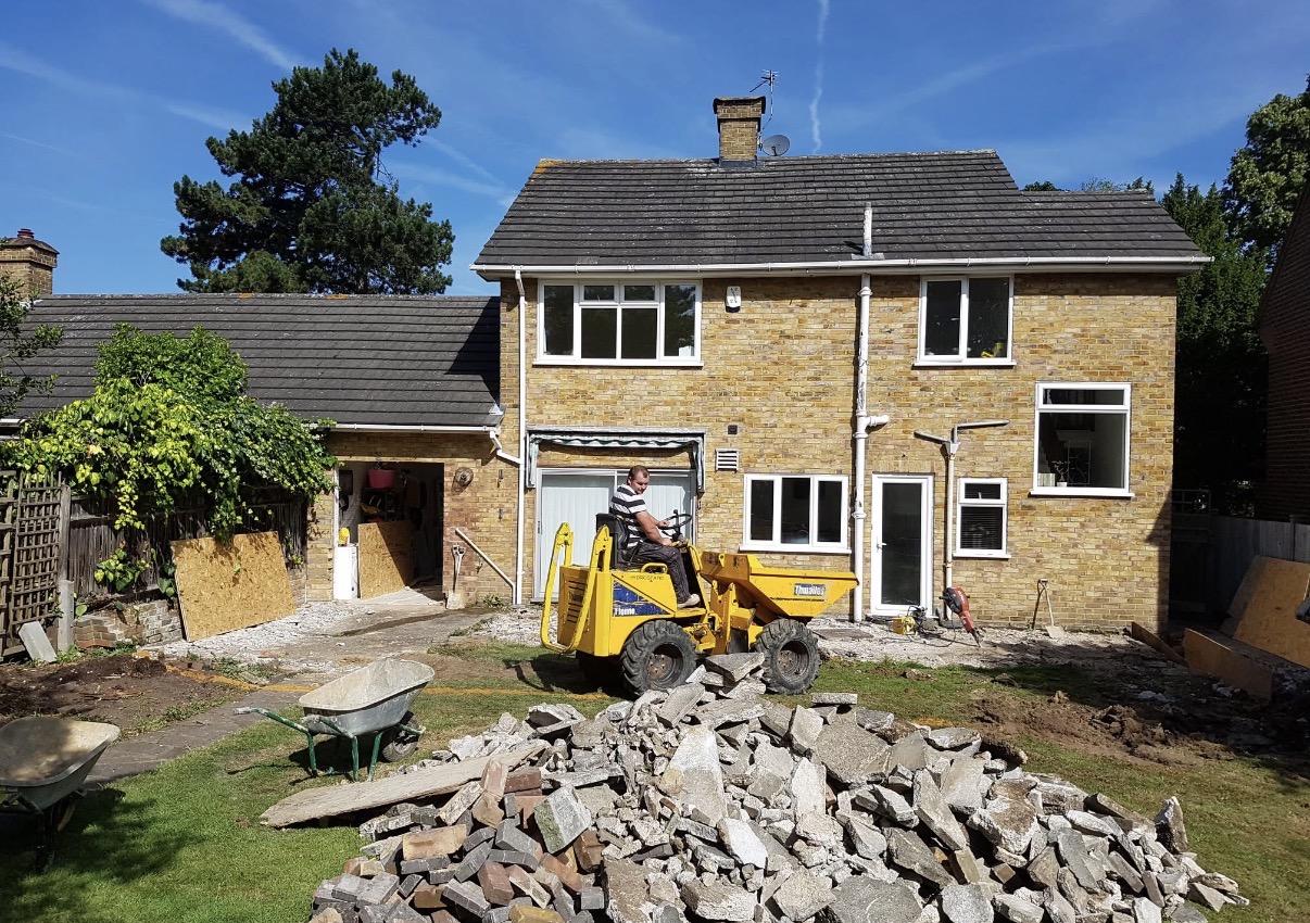 A man uses a mini dumper to clear rubble on a building renovation site in front of a brick house with new windows.