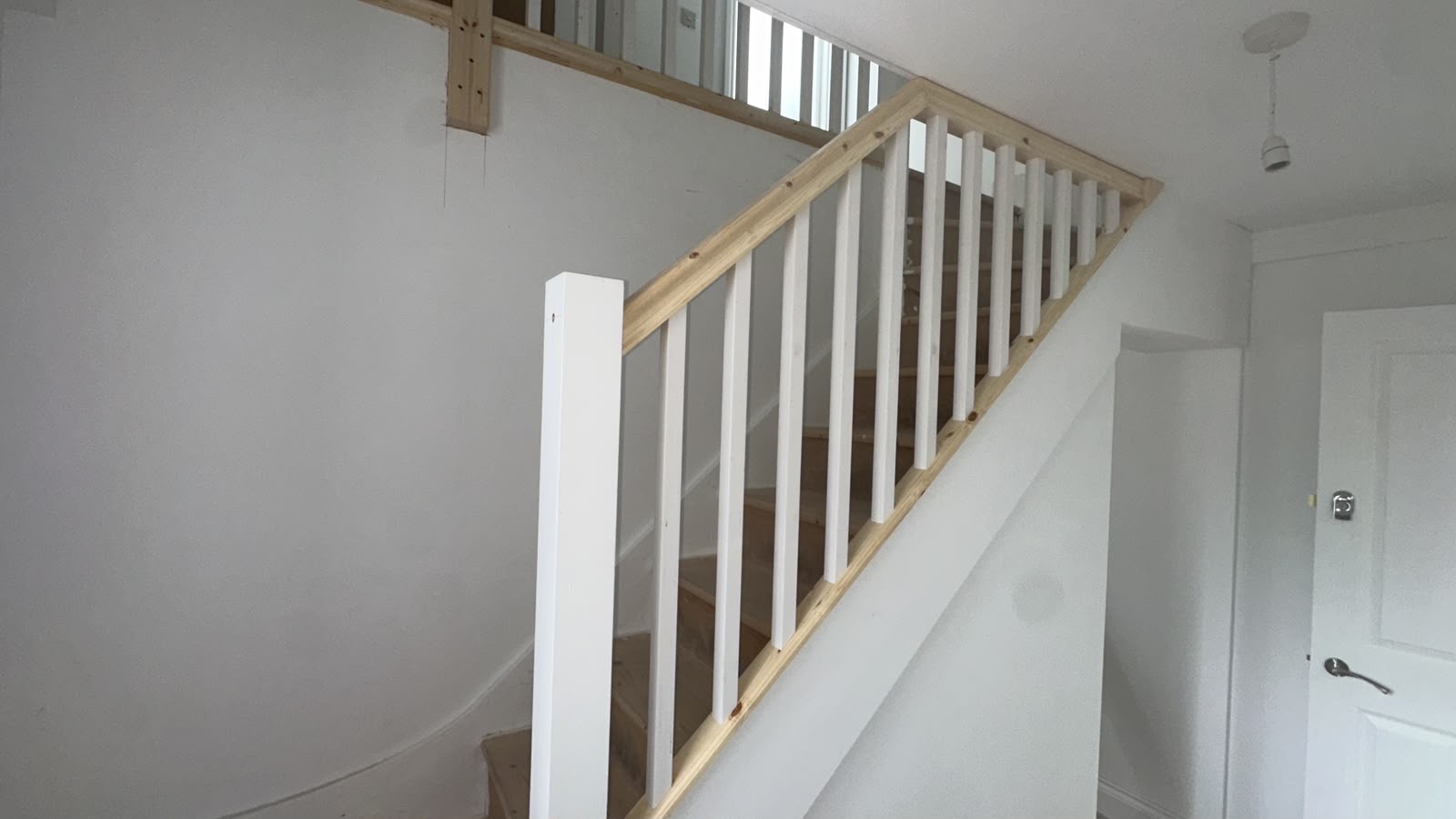 Newly installed wooden staircase balustrade with white spindles and newel post, and an unfinished wooden handrail, in a white hallway.