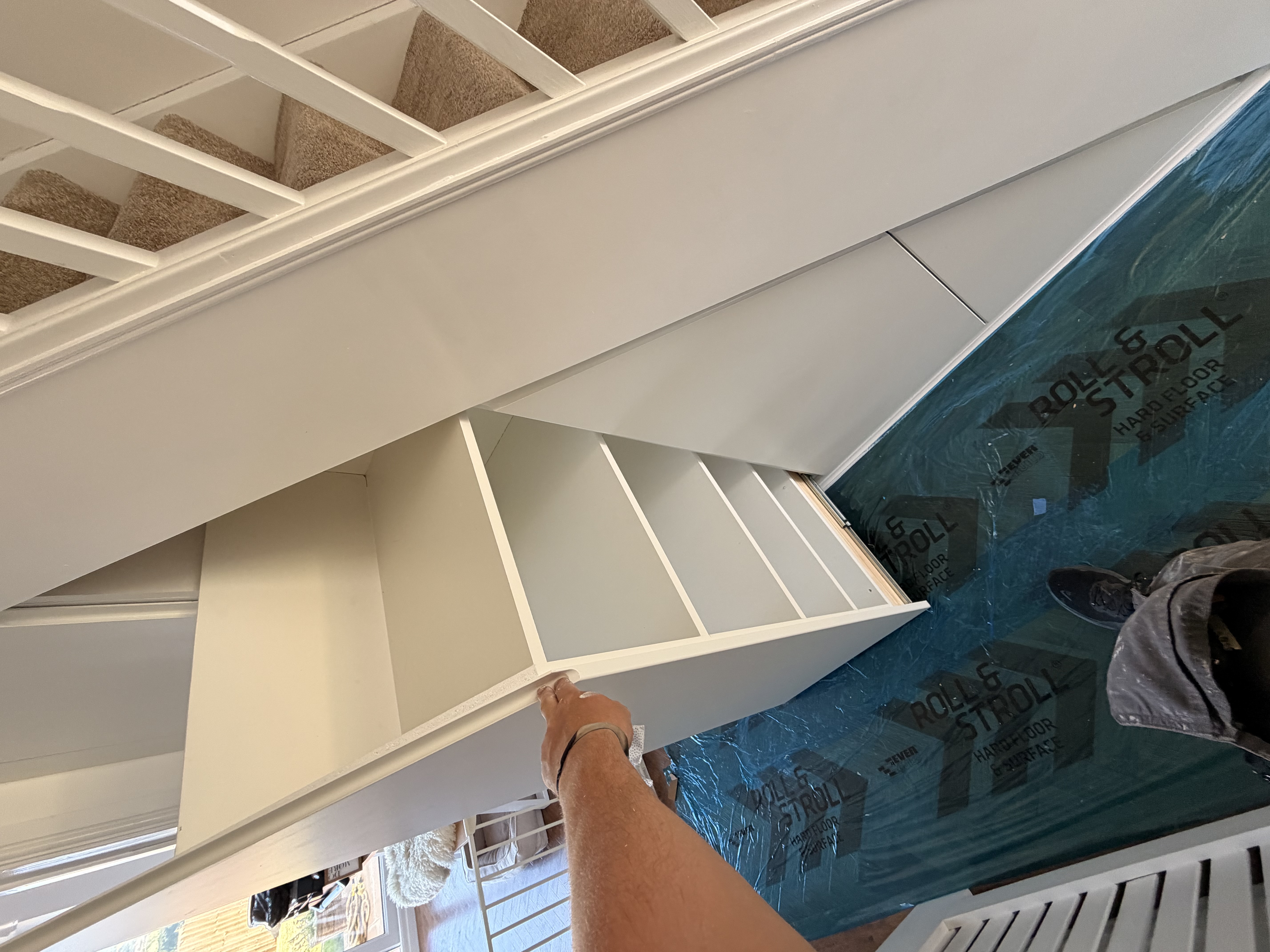 White built-in under-stairs storage unit with shelves being installed next to a carpeted staircase, floor protected.