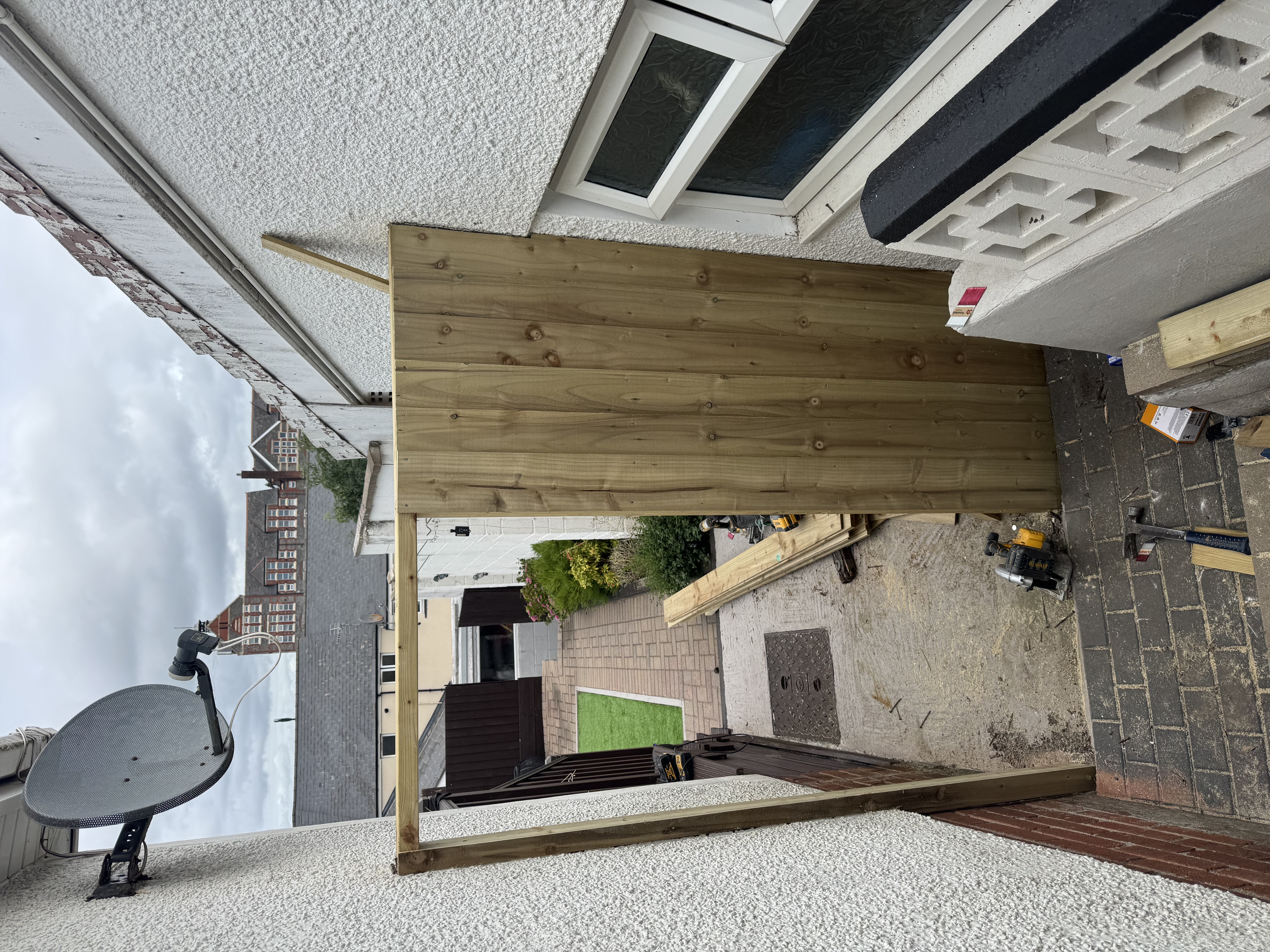 Timber fence panel and frame being installed next to a house wall, with a circular saw and wood debris on the ground.