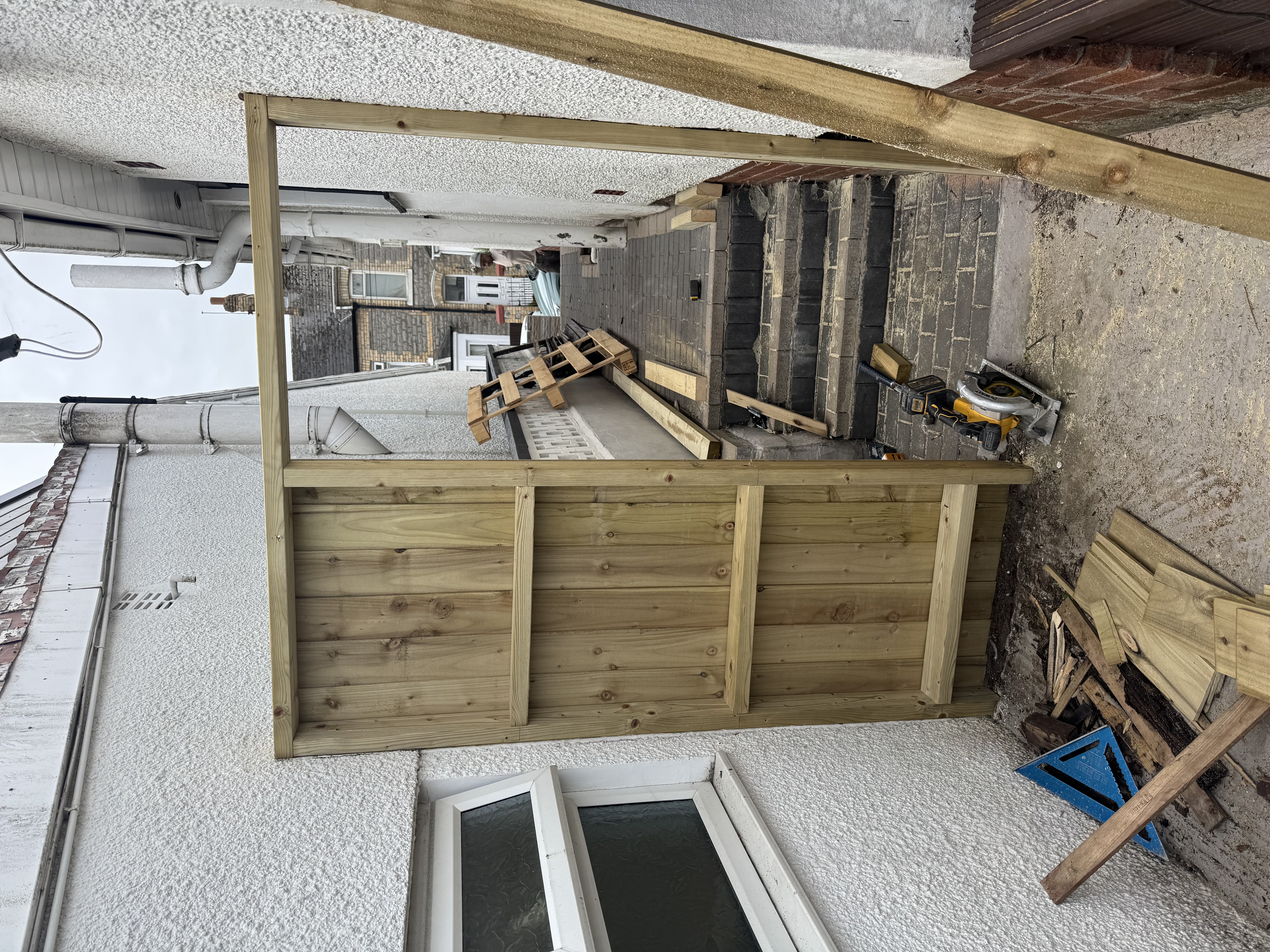 Partially built wooden garden gate and frame in a narrow alleyway with wood offcuts and a circular saw on the ground.