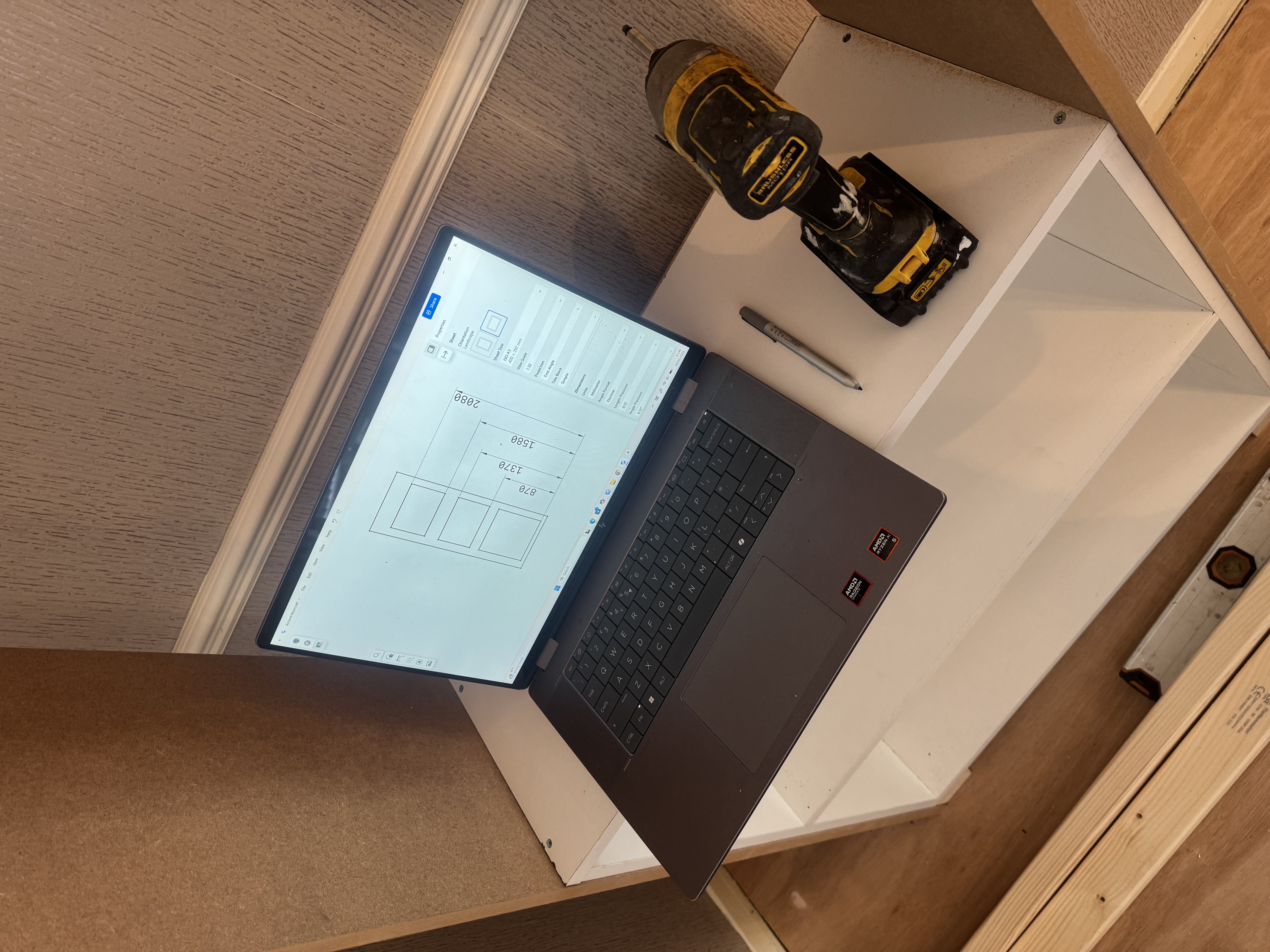 A laptop shows a detailed carpentry design of a door with dimensions, next to a yellow power drill on a white wooden cabinet.