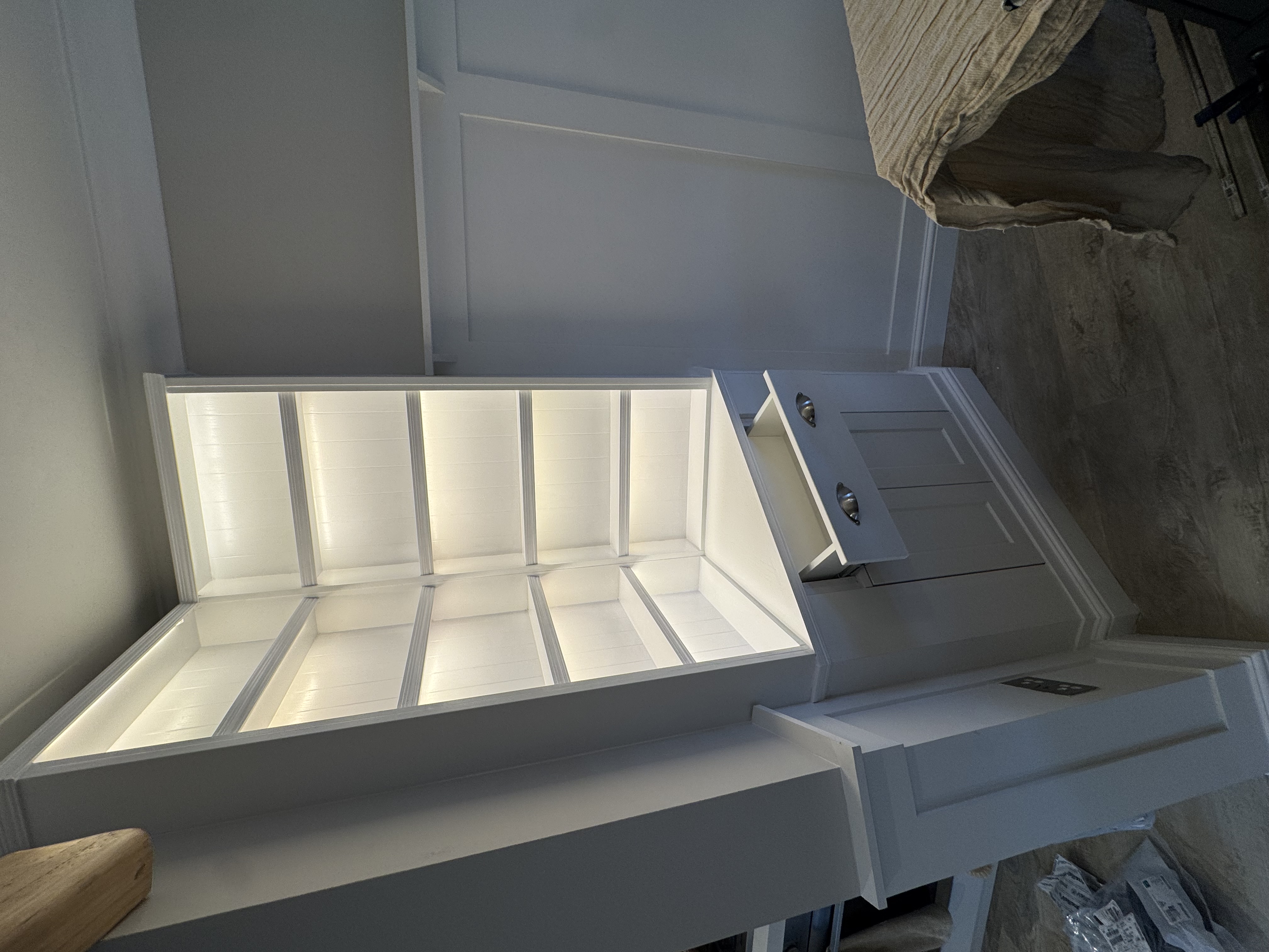 A newly installed white fitted corner cabinet with illuminated shelves, a drawer, and base cupboards, beside matching wall panelling.